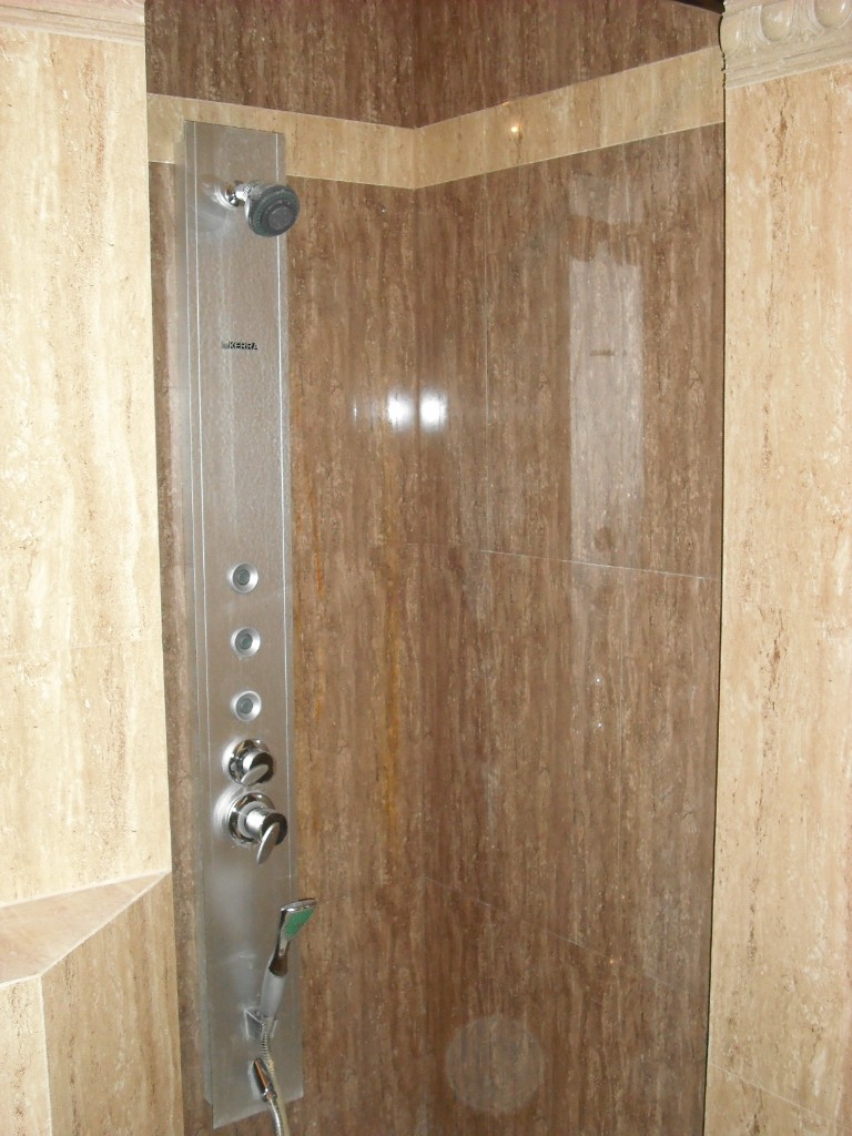 Kabina prysznicowa z panelem natryskowym, wykończona płytkami imitującymi trawertyn w odcieniach beżu i brązu. Panel natryskowy ze stali nierdzewnej z dyszami, baterią i słuchawką prysznicową.